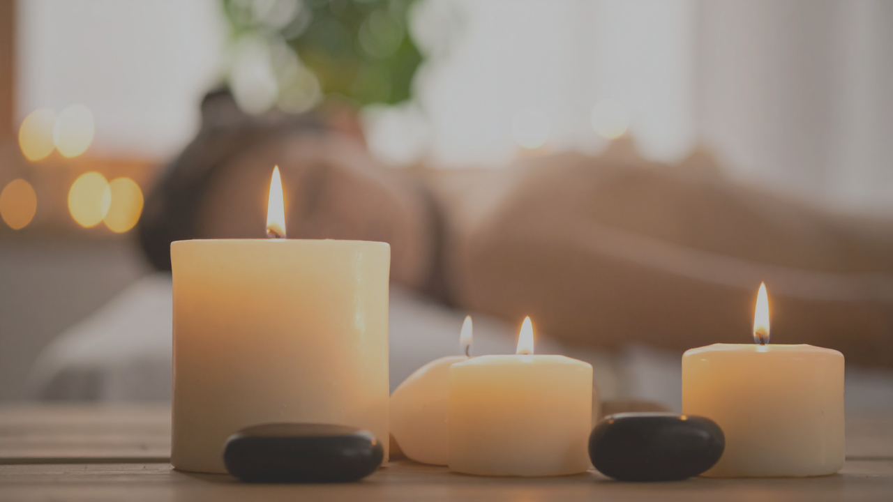 Fotografía en primer plano que captura un ambiente de spa tranquilo, con varias velas de pilar encendidas de color crema y piedras de masaje oscuras y lisas descansando sobre una superficie de madera. La iluminación cálida y tenue crea una atmósfera de paz y bienestar.