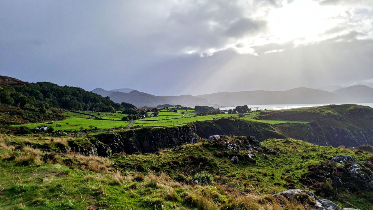 A green Irish coastal landscape with stone walled fields, low hills, and the sea beyond, lit by sun breaking through cloud. Used to accompany the Irish Pagan School blog post 'What Is Irish Paganism?'.