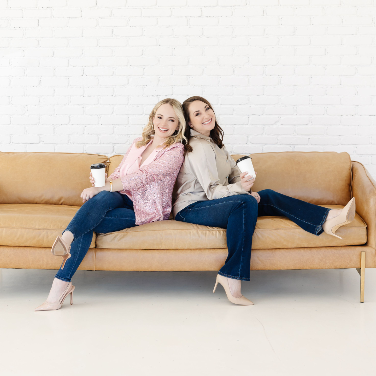 Casey and Nicole sitting back to back on a leather couch, smiling together and holding coffee cups