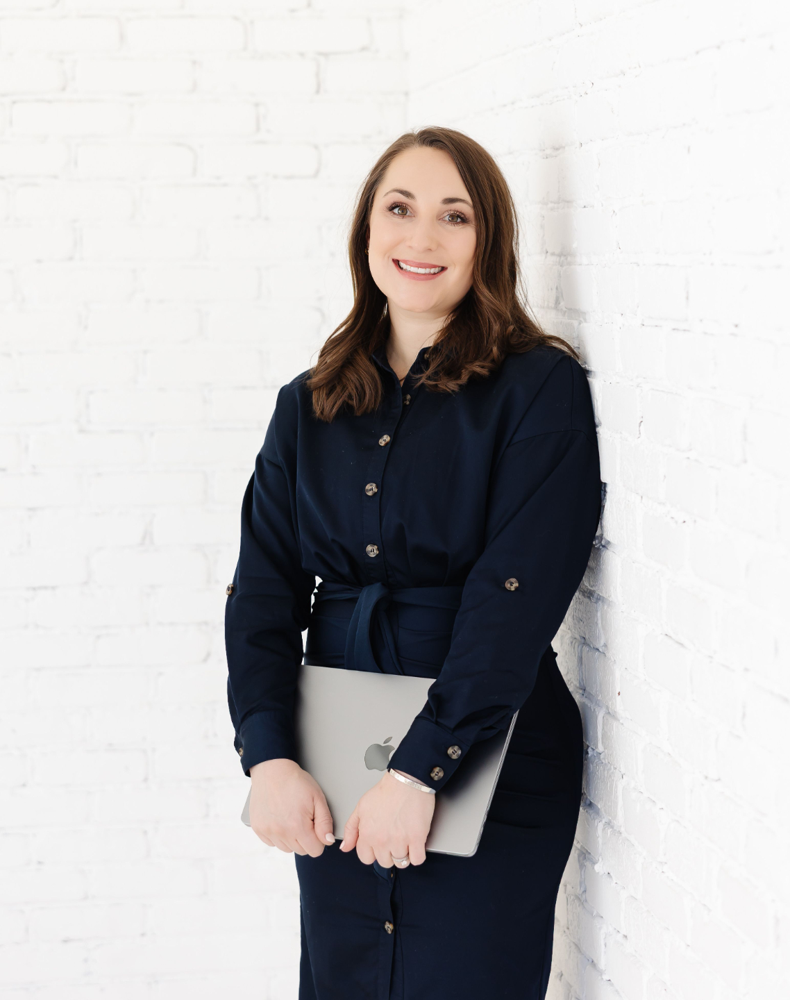 Nicole leaning against a white brick wall, smiling and holding a closed laptop