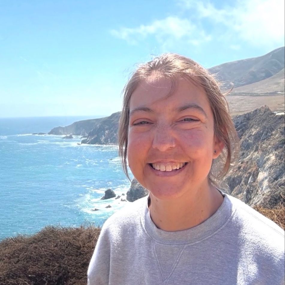a woman is smiling in front of the ocean and a rocky shore behind her