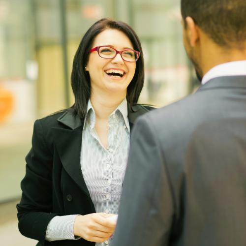 A smiling businesswoman wearing red glasses and a black blazer laughs during a conversation with a man in a suit. The two professionals appear to be networking or having a friendly workplace discussion in a bright office or conference setting, suggesting positive communication, employee engagement, professional connection, and relationship-building.