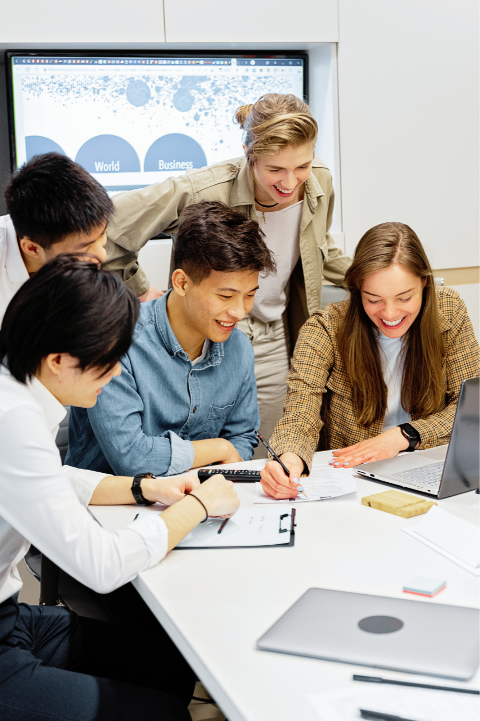 A diverse group of five smiling professionals collaborates around a conference table during a workplace training session. One woman writes on a printed worksheet while others lean in and look on, with laptops, papers, a calculator, and office supplies on the table. A presentation screen in the background shows business-related graphics, suggesting an interactive continuing education, team learning, or employee development workshop.