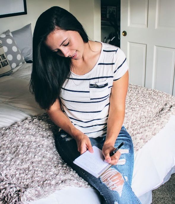 A woman with long dark hair sits on the edge of a neatly made bed, looking down as she writes in a small spiral notebook with a pen. She is wearing a white short-sleeve shirt with thin navy horizontal stripes and ripped blue jeans. A textured gray blanket is spread across the bed beside her, with pillows and neutral bedroom decor in the background. The image has a calm, reflective, personal feel, suggesting journaling, self-care, planning, or quiet reflection at home.