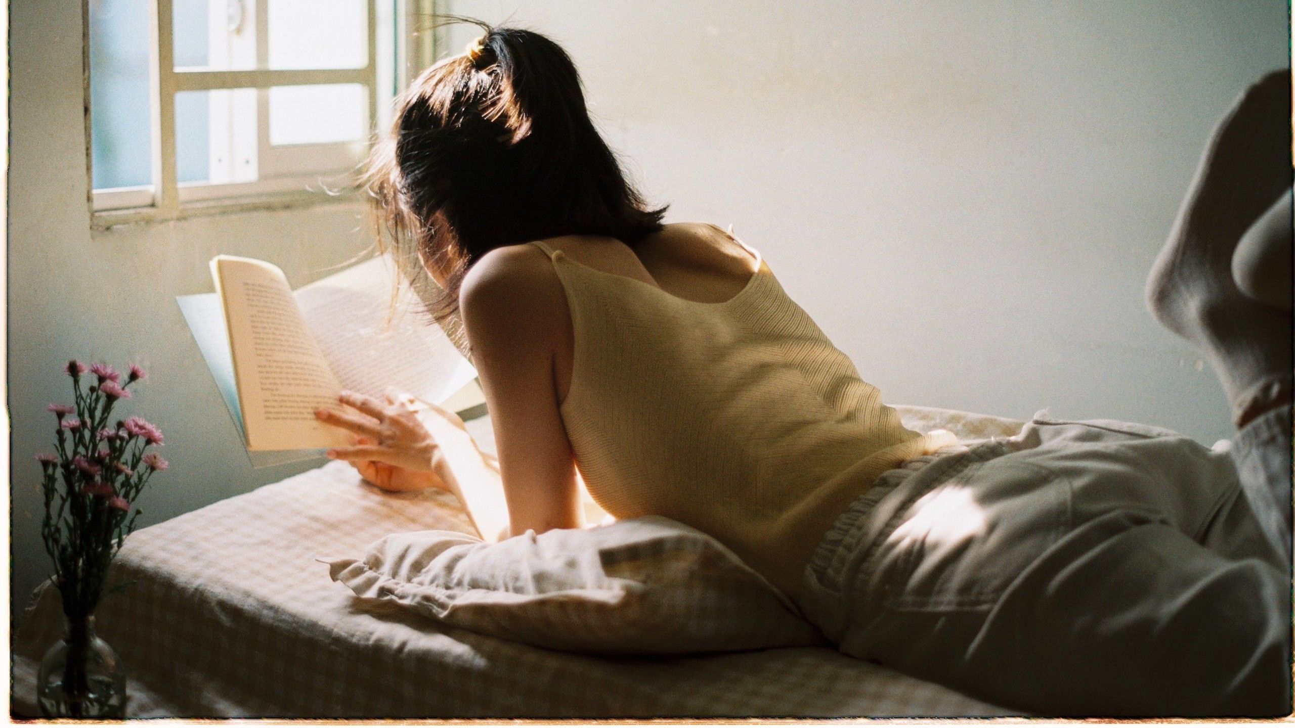 A woman lies on her stomach reading a book by the window, enjoying a peaceful afternoon.