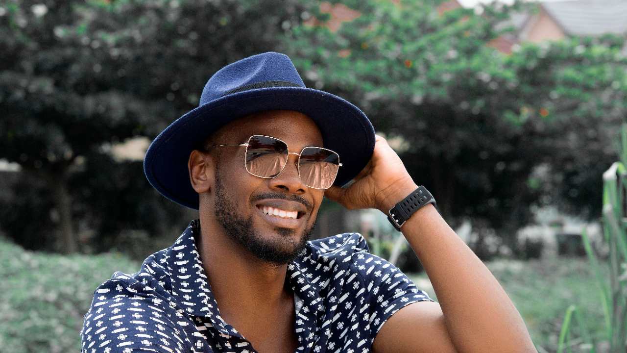 Portrait of a fashionable man with a blue hat and sunglasses, smiling outdoors.