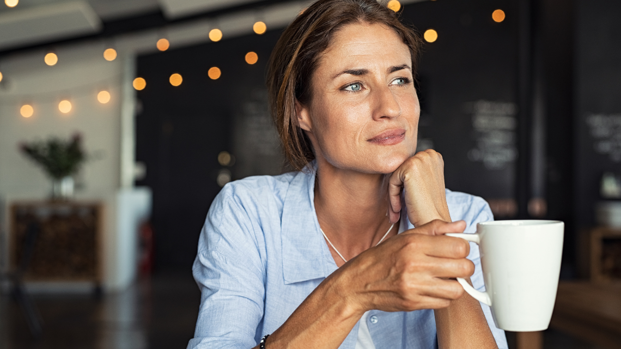 Woman in a cafe resting her chin on her hand, looking thoughtfully into the distance beside a cup of coffee.
