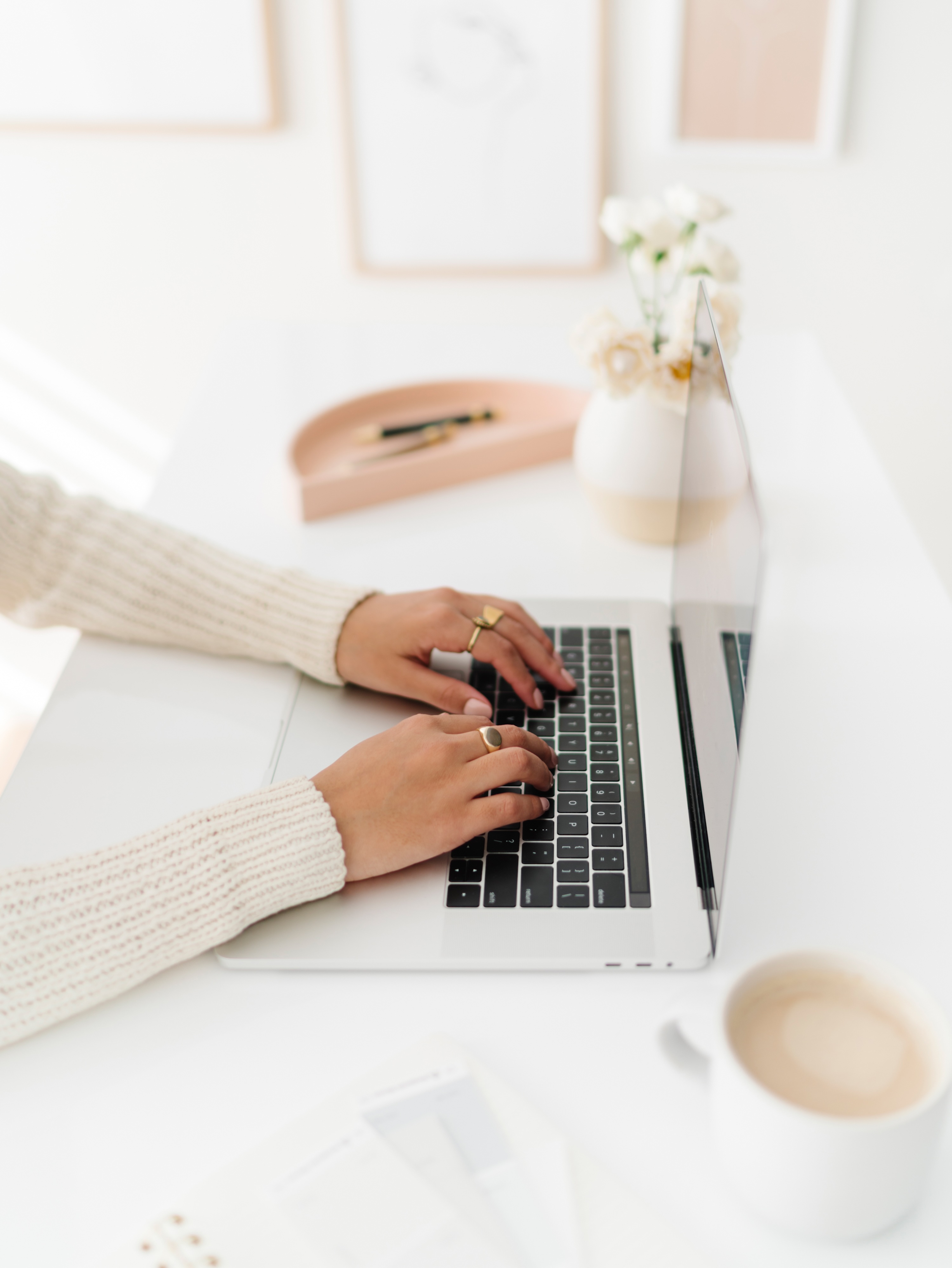 Hands in a cream cardigan typing on a laptop beside coffee and white flowers