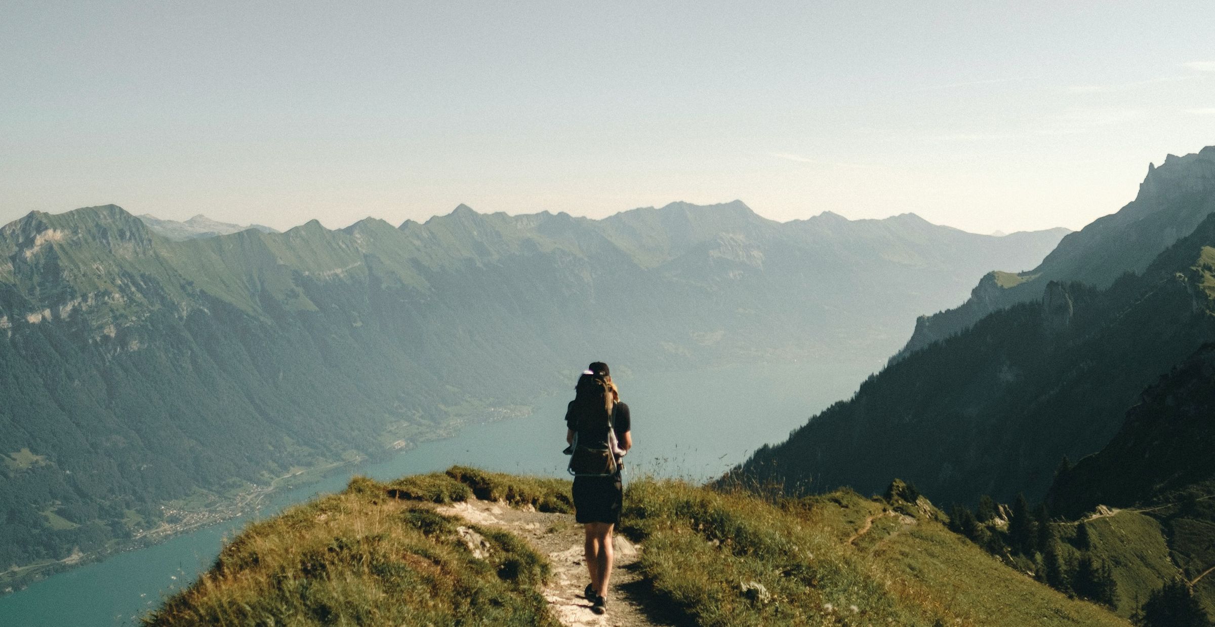 Background Image: Hiker walking away from camera on a mountaintop trail.  They are looking at a river valley with mountains on either side