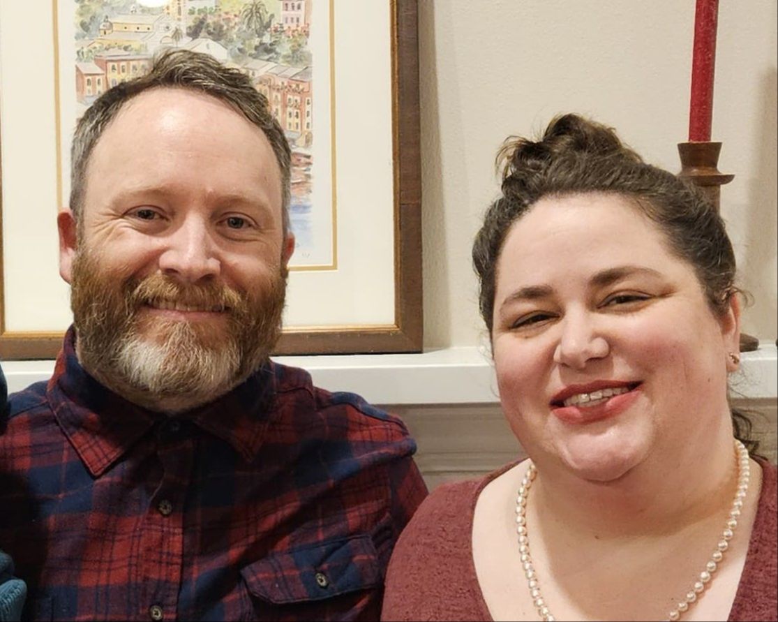 Headshots of a man and a woman smiling at the camera with a painting of buildings and a candle in the background