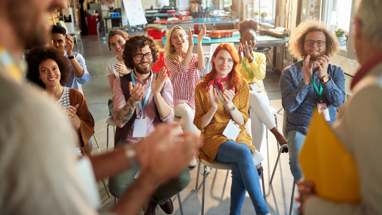 A diverse group of teachers and employees seated in a bright, modern training space applauds an off-camera presenter during an interactive professional development session. Participants wear conference badges and appear engaged, smiling, and enthusiastic as they take part in collaborative workplace and educator training.