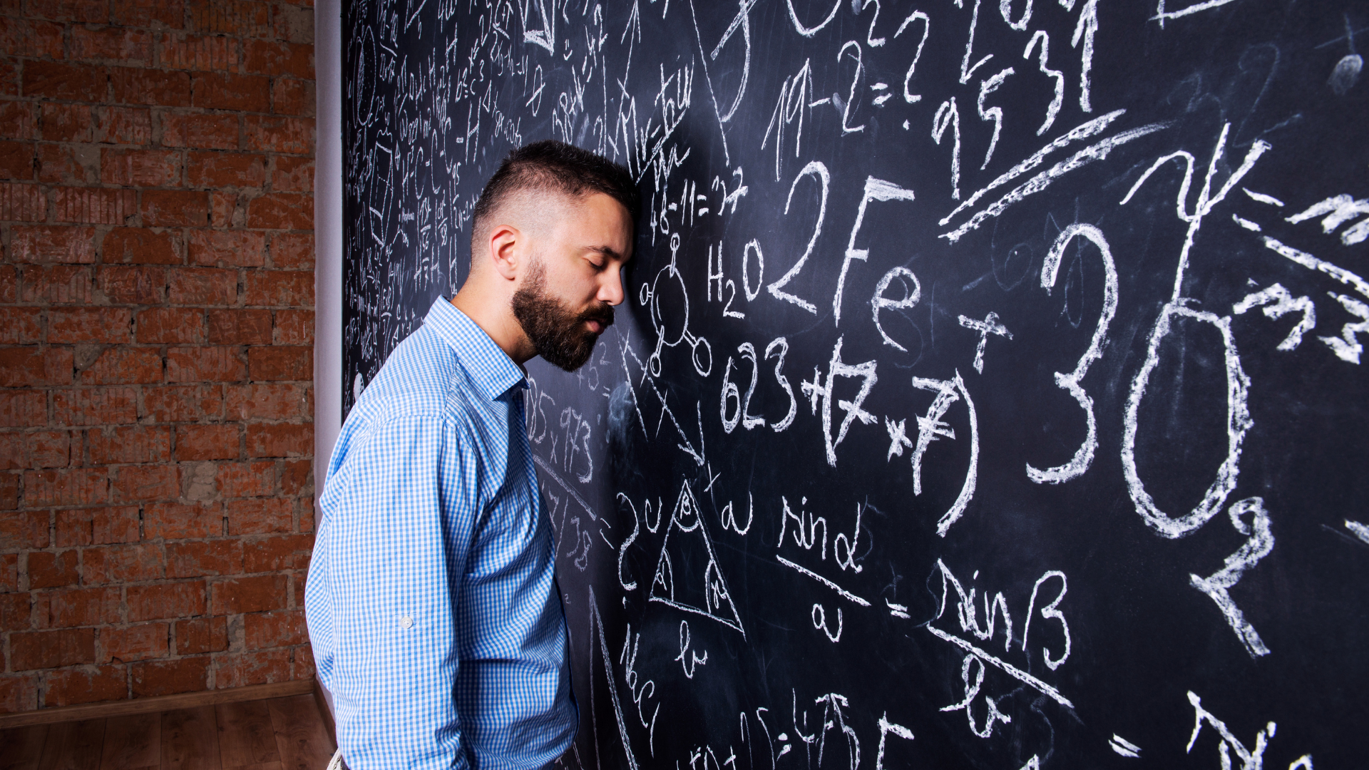 A visibly exhausted teacher in a blue checkered button-down shirt leans his head against a large black chalkboard covered in complicated math equations, formulas, numbers, and scientific symbols written in white chalk. His eyes are closed, and his posture suggests stress, overwhelm, and mental fatigue. An exposed red brick wall and wood flooring give the setting an industrial classroom or training-room feel. The image represents teacher mental health, educator burnout, emotional exhaustion, workplace stress in schools, and the pressure teachers can feel while managing complex responsibilities.