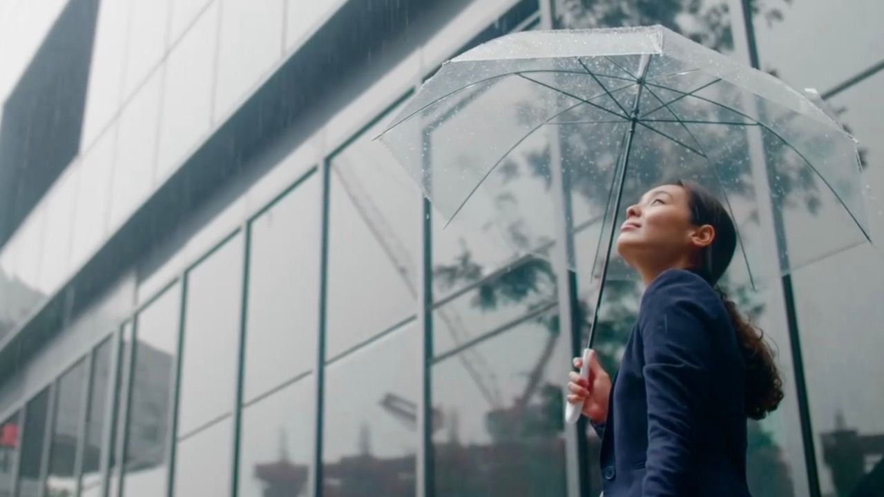 A woman in a navy blazer stands outside a modern glass building on a rainy day, holding a clear umbrella above her head. She looks upward with a calm, reflective expression as raindrops fall around her. The image represents emotional strength, adaptability, and the ability to stay grounded during difficult moments, making it a strong visual for a resilience course.
