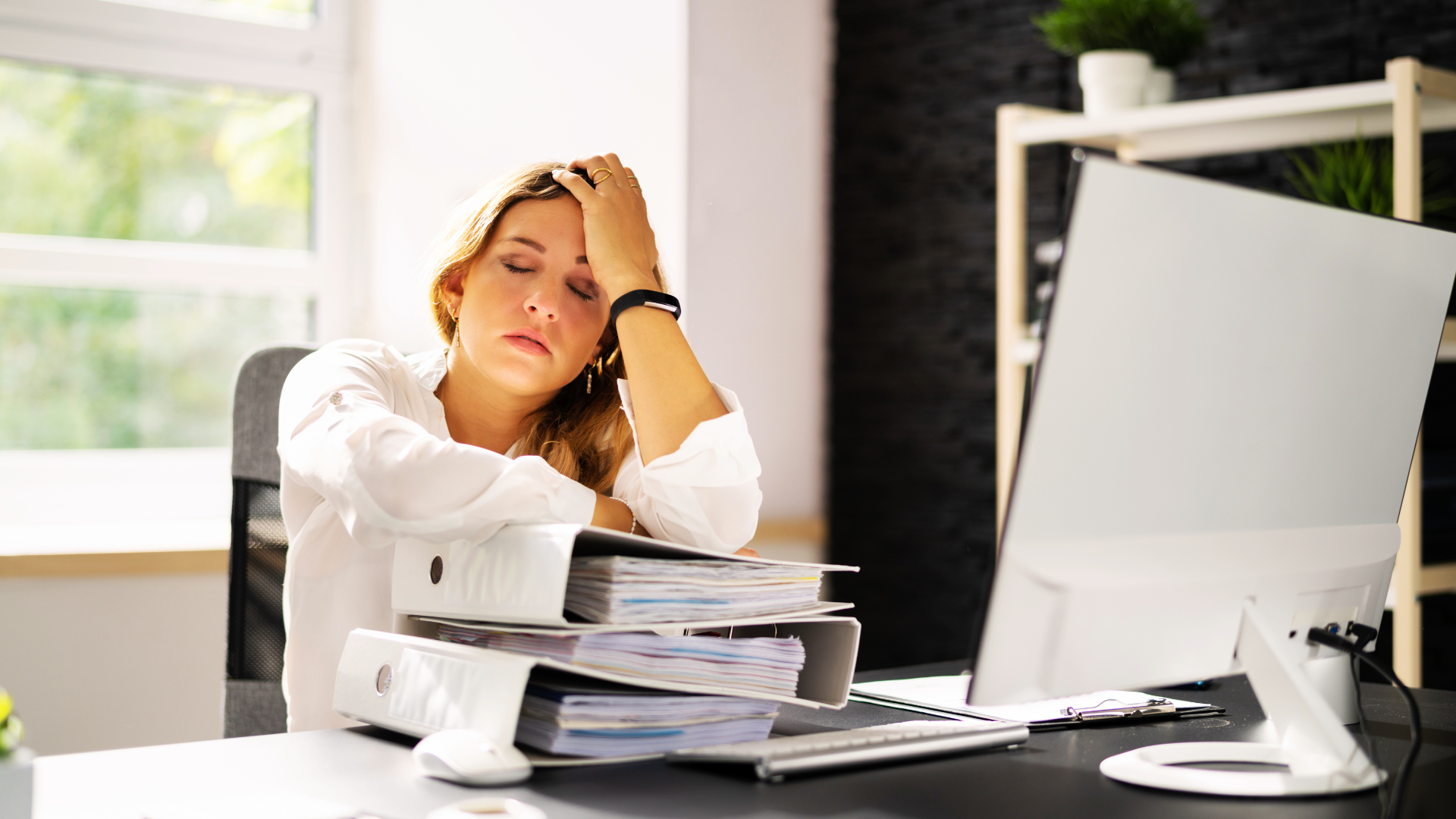 A visibly exhausted employee sits at a desk in a bright office, resting her head in one hand with her eyes closed. In front of her are two overfilled binders stacked with papers, a keyboard, mouse, clipboard, and a large computer monitor. Sunlight streams in from a window behind her, while a shelf with small plants appears in the background. The image conveys workplace stress, burnout, fatigue, overwhelm, and the need for employee mental health support.