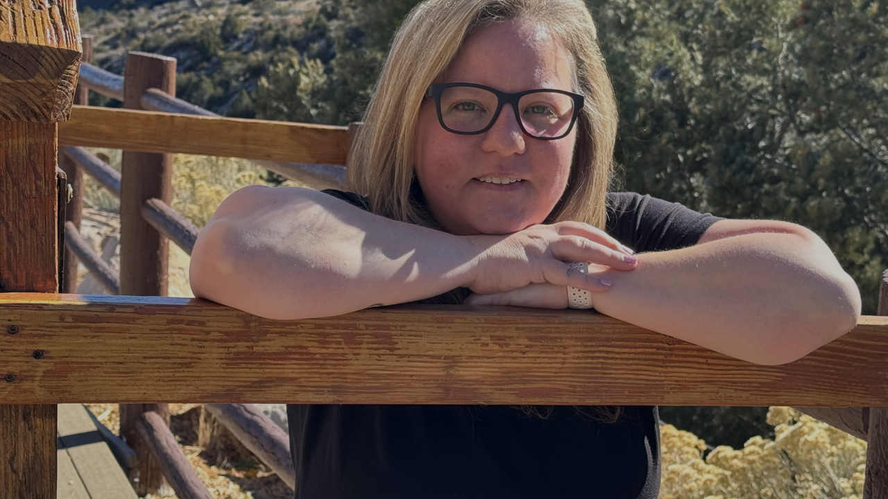 Image of Melinda in the mountains with a brown and pink jacket with blue jeans and brown boots. Snow on the ground and pine trees behind her.