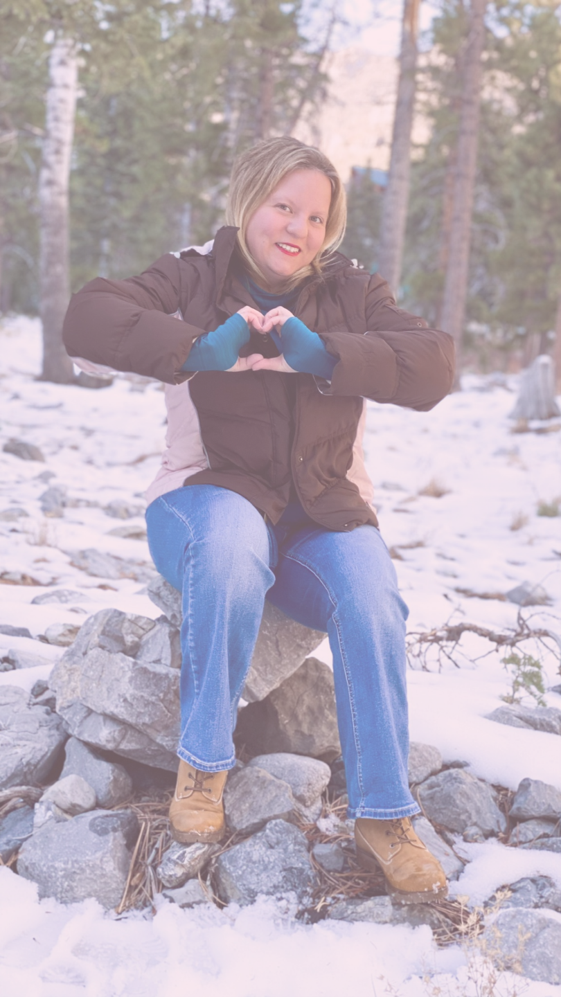 Melinda in the snowy mountains with trees in the background and snow on the ground. She is sitting on rocks and putting her hands in a heart. She is wearing a brown jacket and blue jeans.