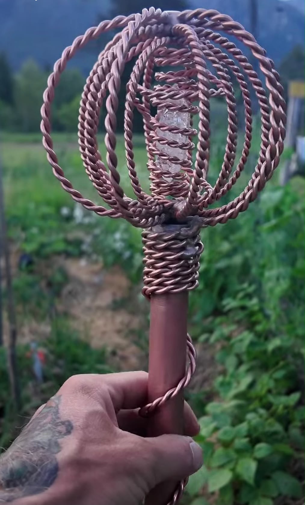 A hand-forged Homestead Harmonizer Antenna System in hand, its crown and quartz heart against the mountains