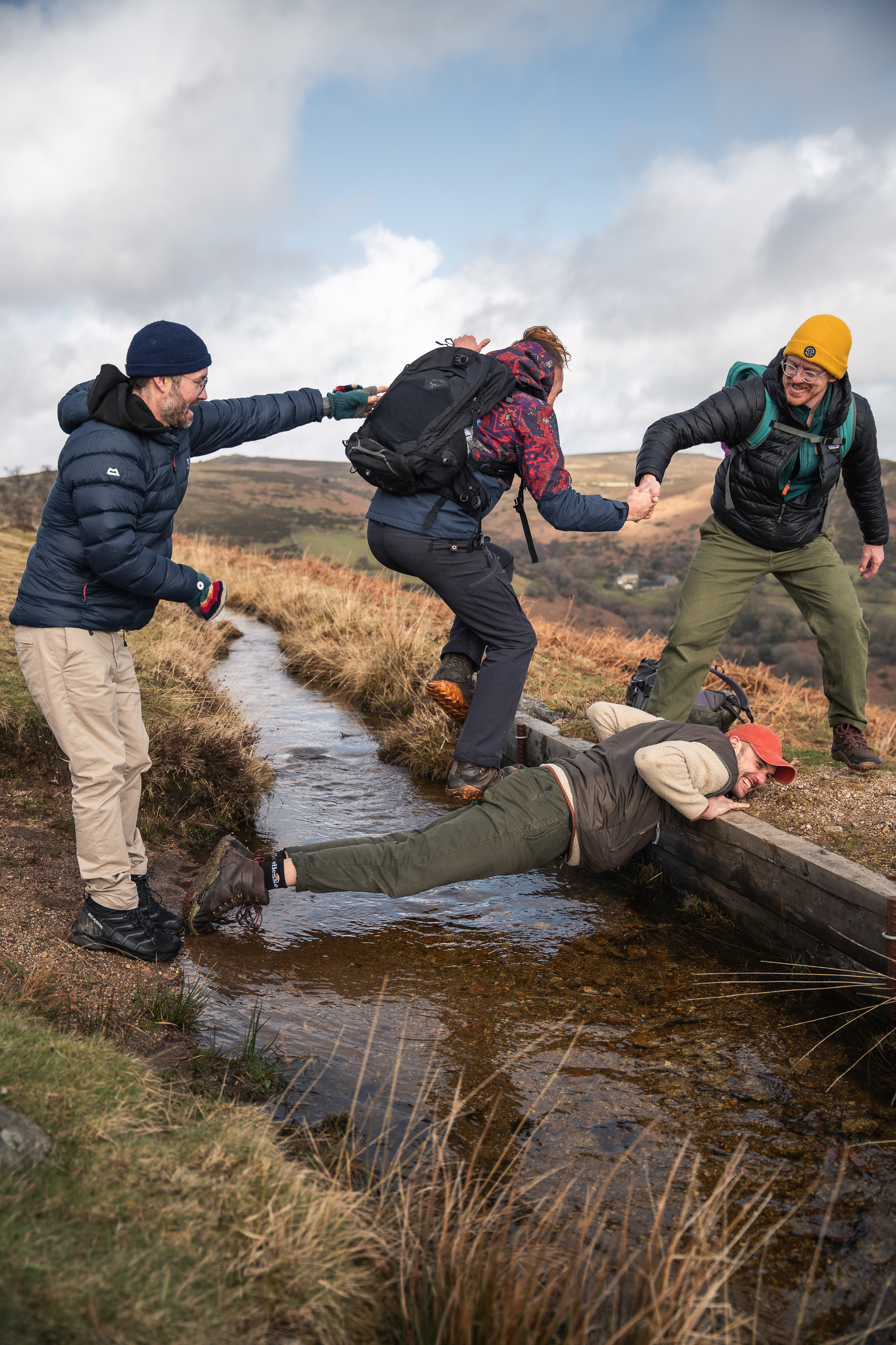 Men of Earth hikers laughing as they help each other across a moorland stream