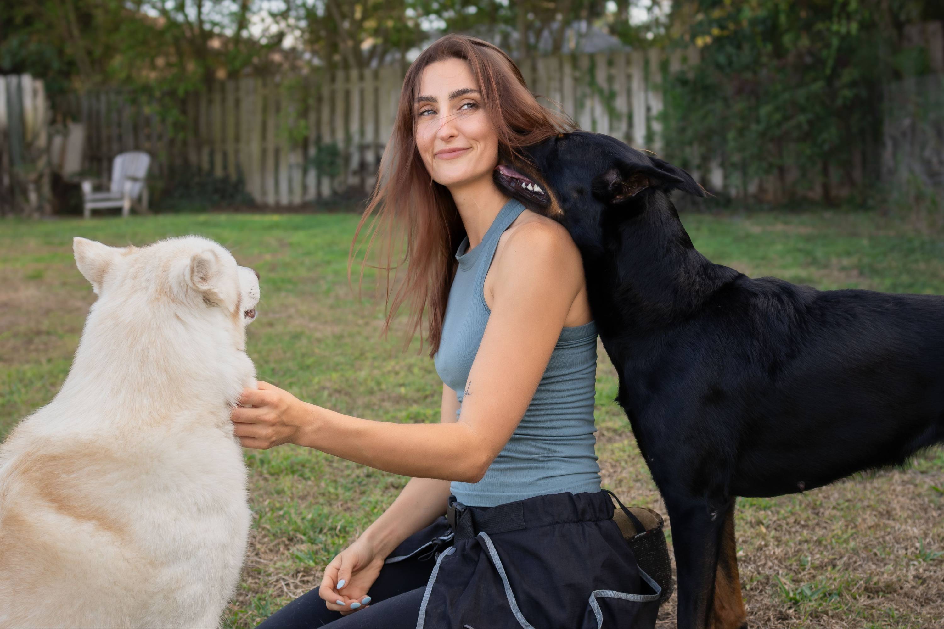 Savanah kneeling in a backyard between a white dog and a black dog during a training session