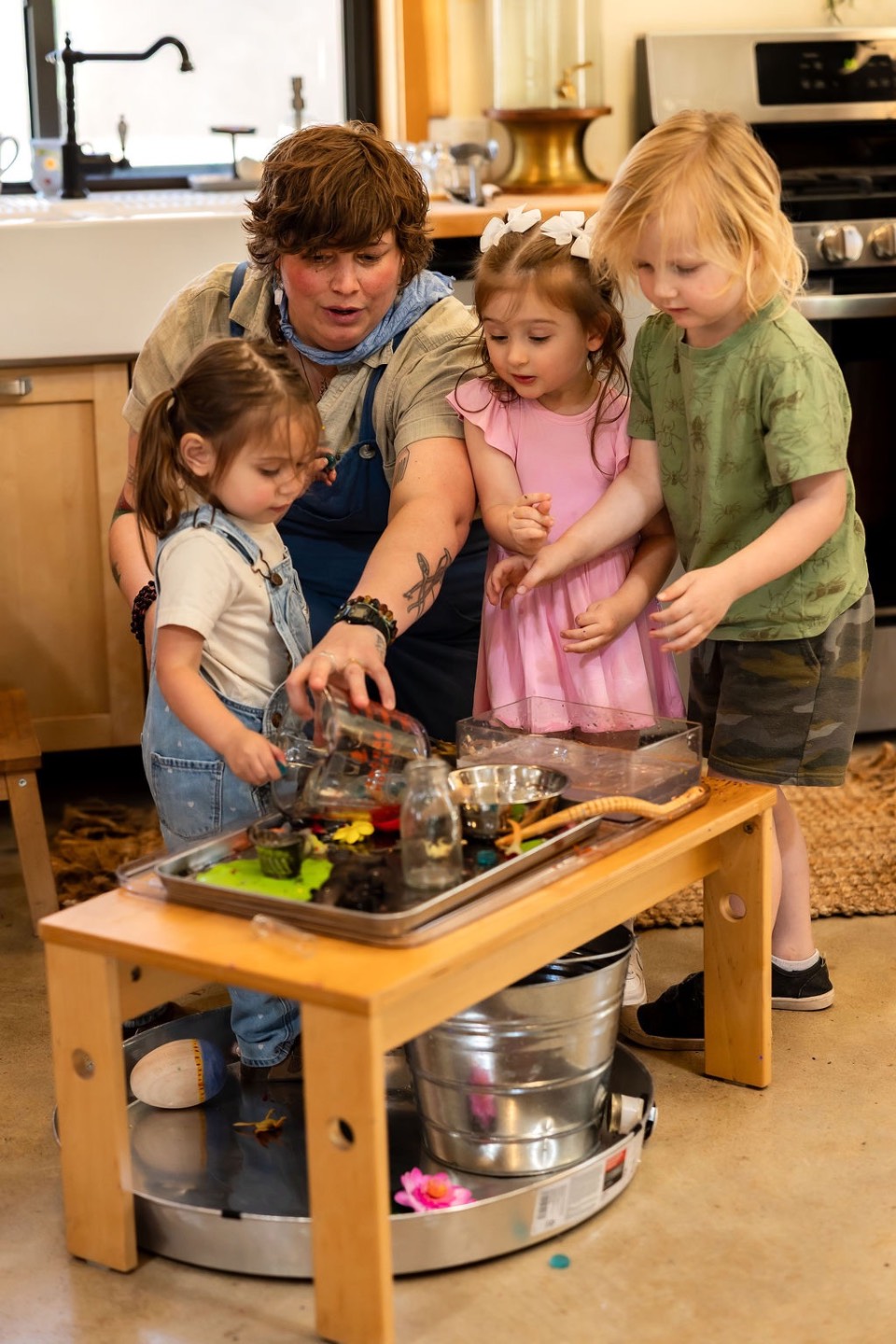 A teacher helping three young children pour and scoop at a water table