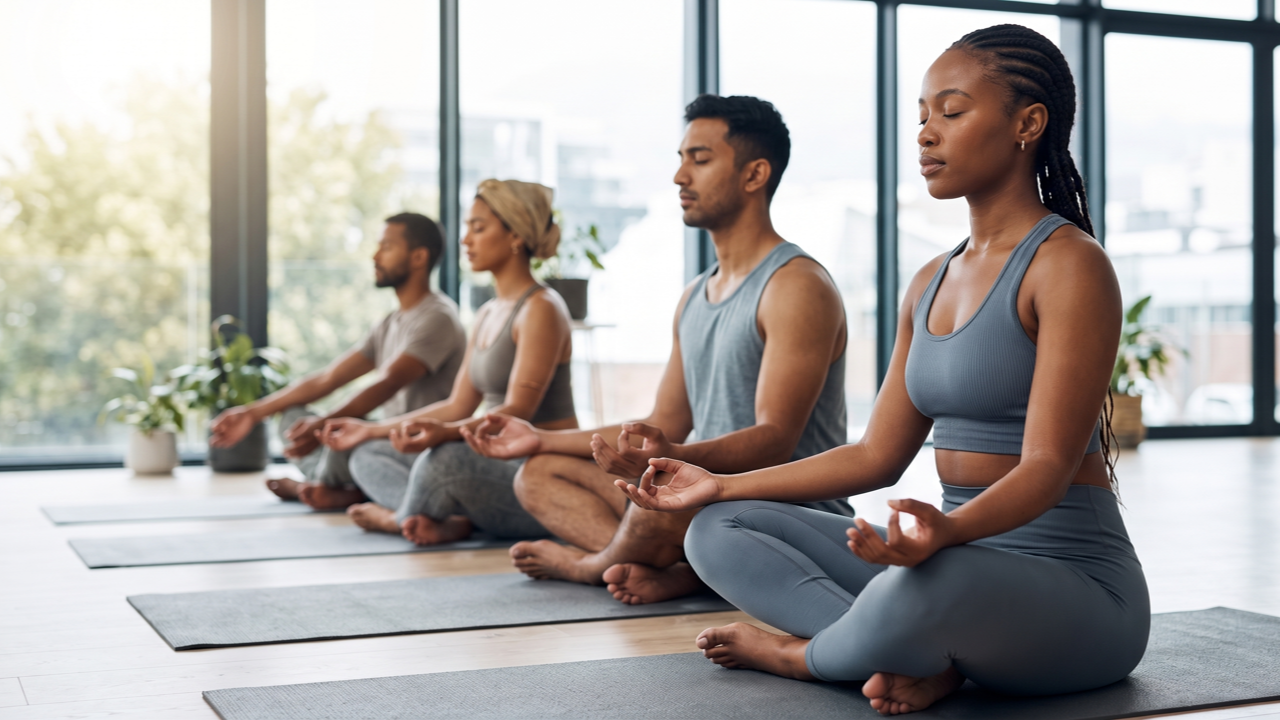 Four students of various ethnicities sit in a row on yoga mats, eyes closed in deep meditation against a backdrop of large windows and city views.