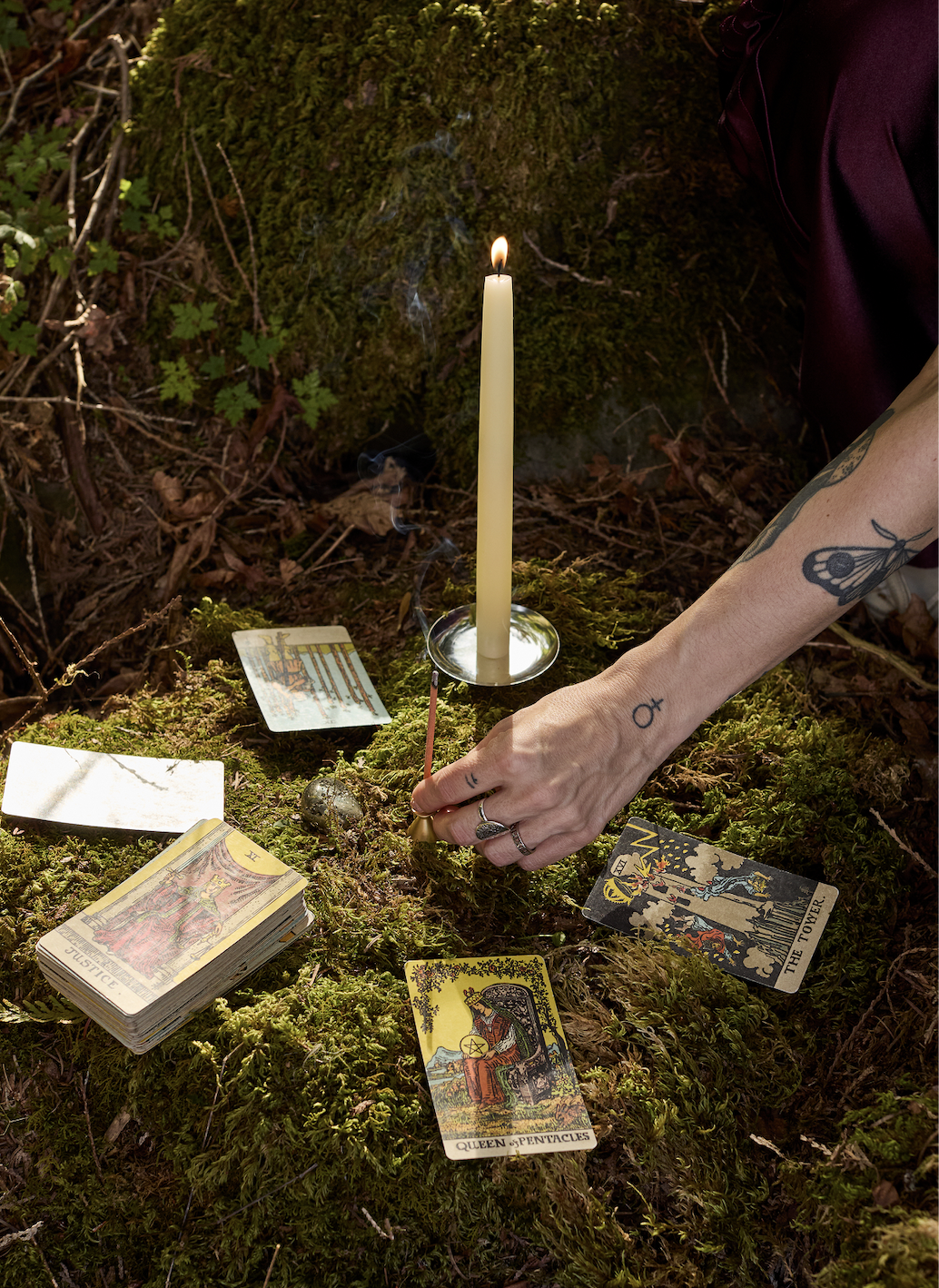 Une main allume un bâton d'encens près d'une bougie, entourée de cartes de tarot sur la mousse
