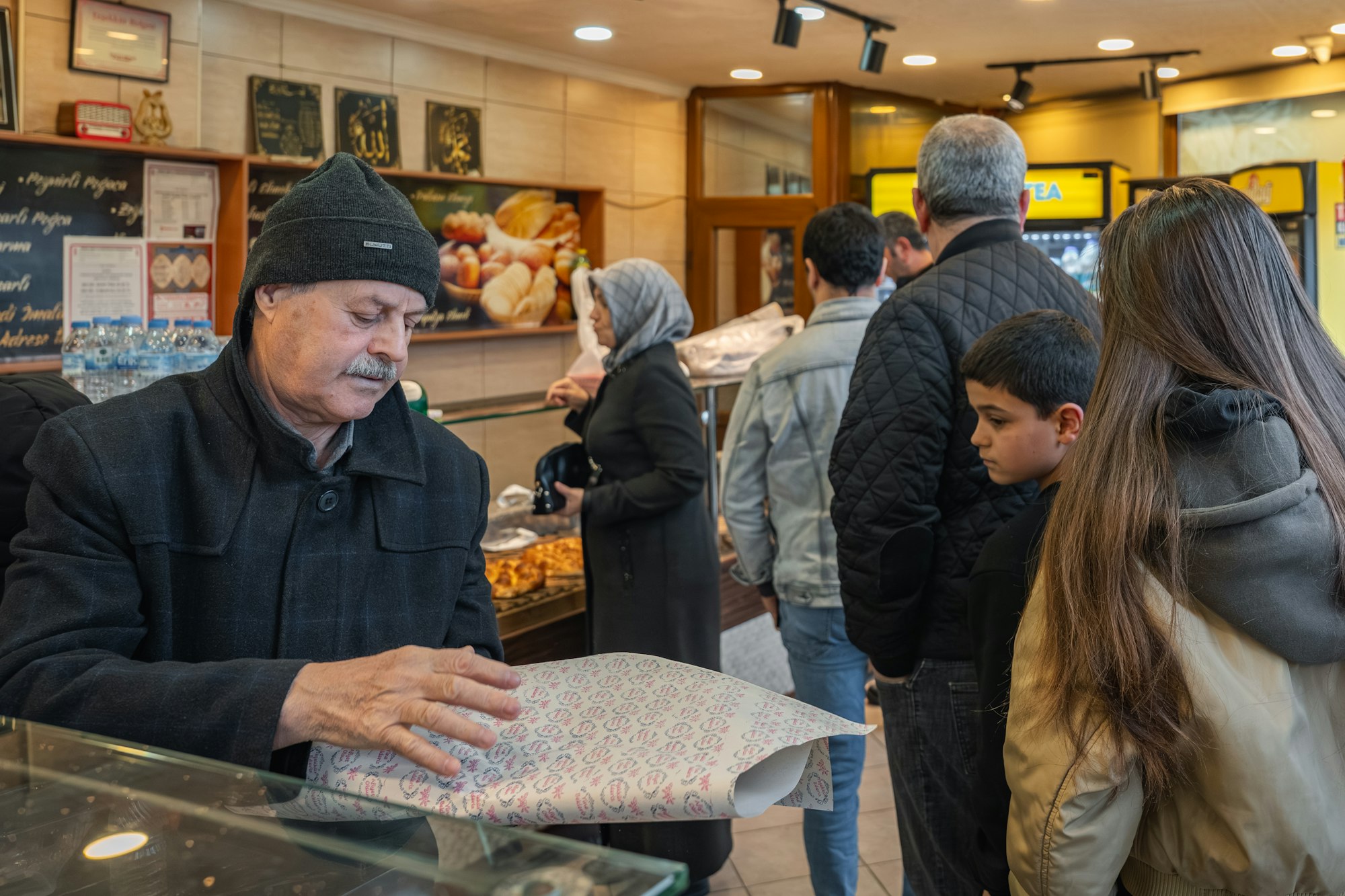 A baker wraps pastries at a counter while customers wait their turn