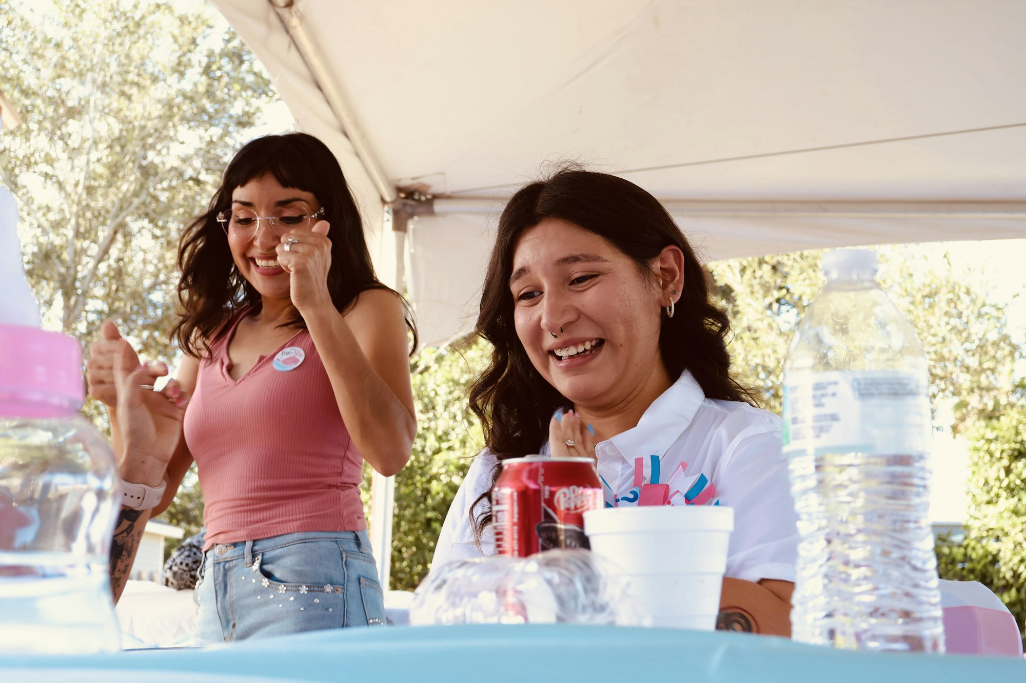 Two women laugh together at a table under a tent