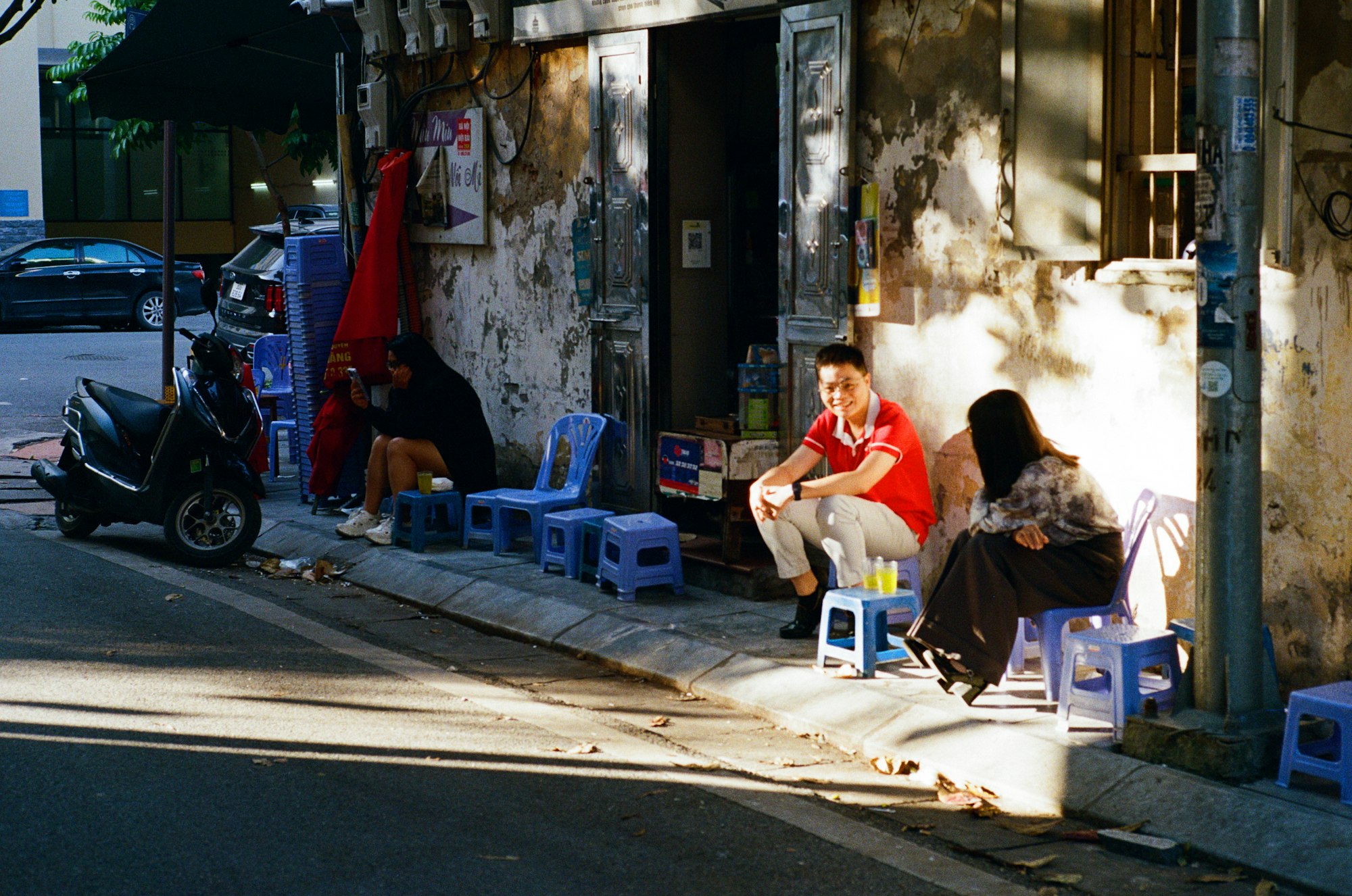A man and a woman talking quietly on low stools along a sunlit street