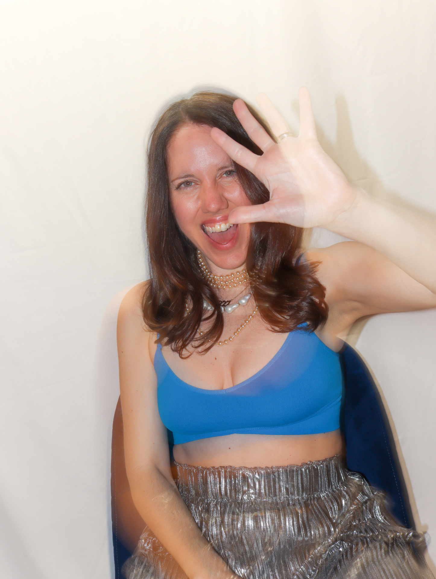 Tamra laughing with one hand raised toward the camera, in a blue top, pearls and a silver pleated skirt