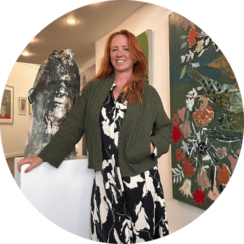 Photo of a woman standing in a gallery space beside artwork, smiling warmly with paintings and a sculpture displayed around her.
