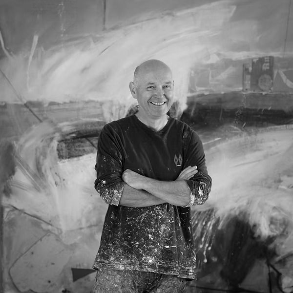 Black and white portrait of artist David standing in his studio, smiling with arms crossed in front of a large expressive abstract painting, wearing a paint-splattered shirt.