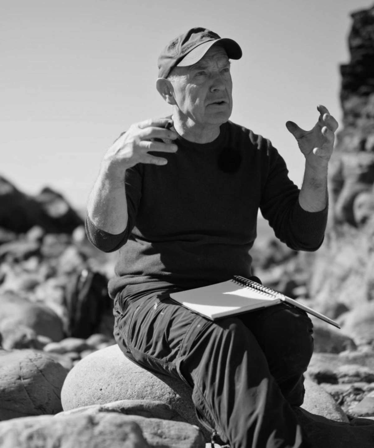 Black and white photo of David sitting on rocks outdoors, gesturing expressively as he speaks, with a sketchbook on his lap in a natural landscape setting.