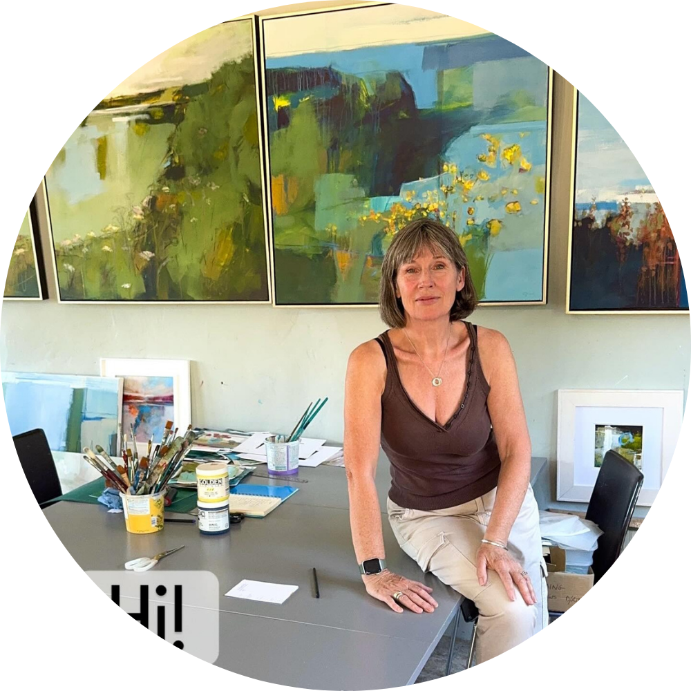 Photo of a female artist sitting at her studio desk, surrounded by paintbrushes and abstract paintings, in a bright creative workspace.