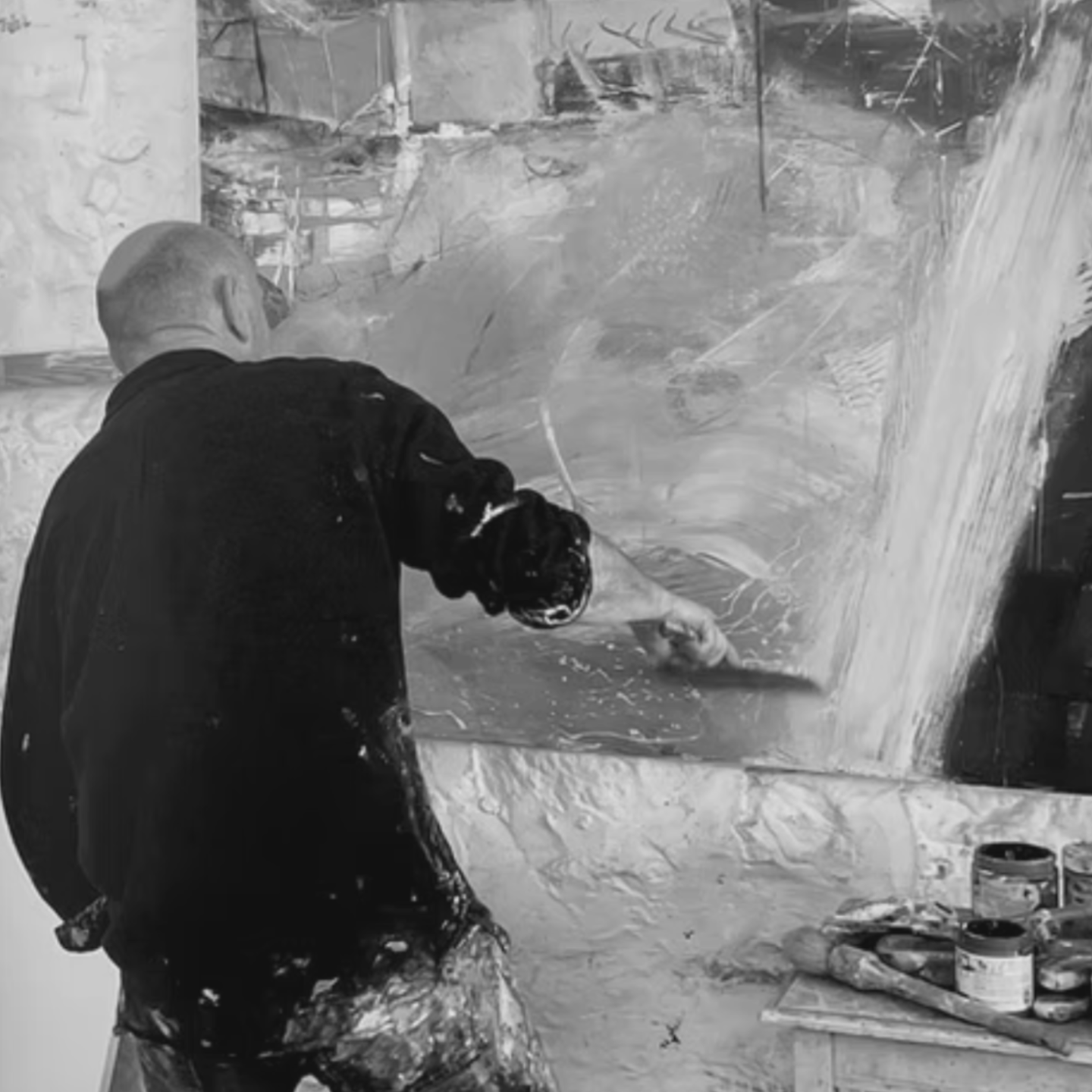 Black and white photo of David painting energetically on a large canvas in his studio, using expressive brushstrokes with paint-splattered clothing.