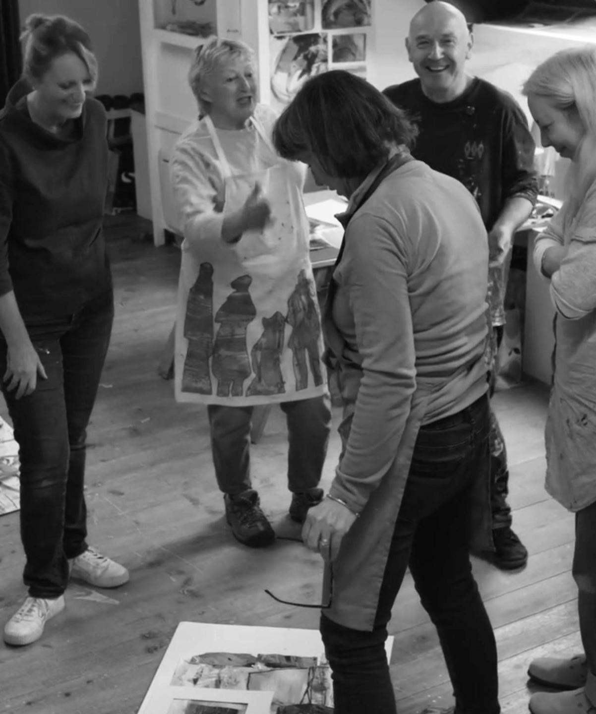 Black and white photo of David and a group of students in a studio, gathered around artwork on the floor, sharing feedback and engaging in a collaborative creative session.