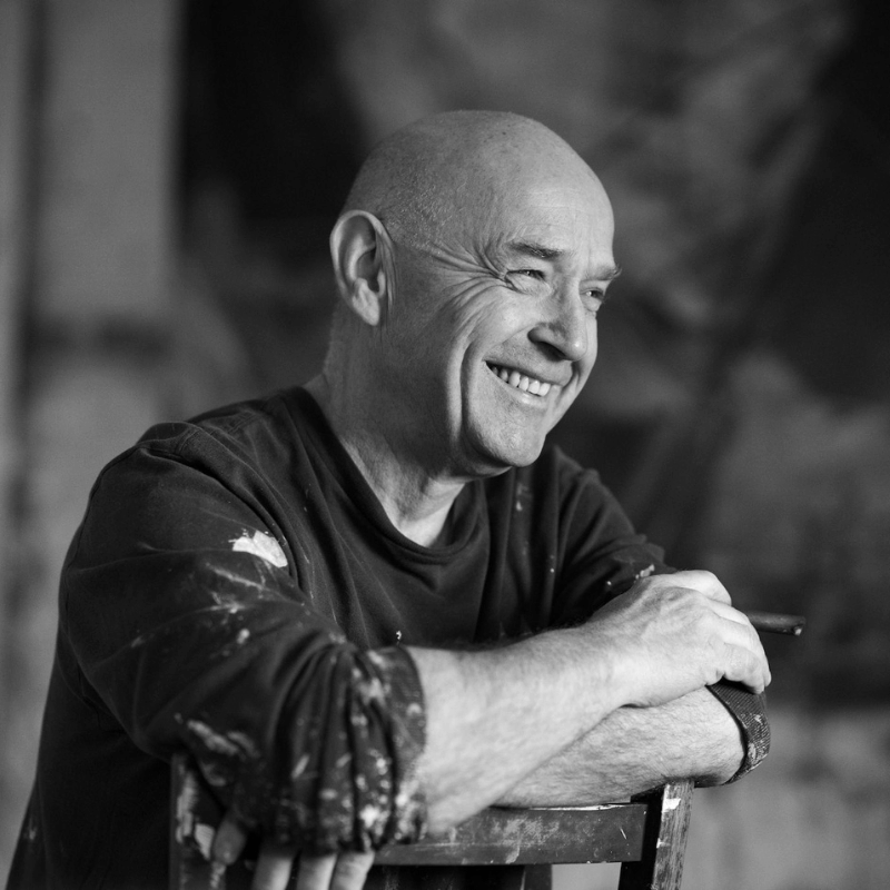 Black and white portrait of artist David Mankin smiling in his painting studio, wearing a paint-splattered shirt and holding a paintbrush.