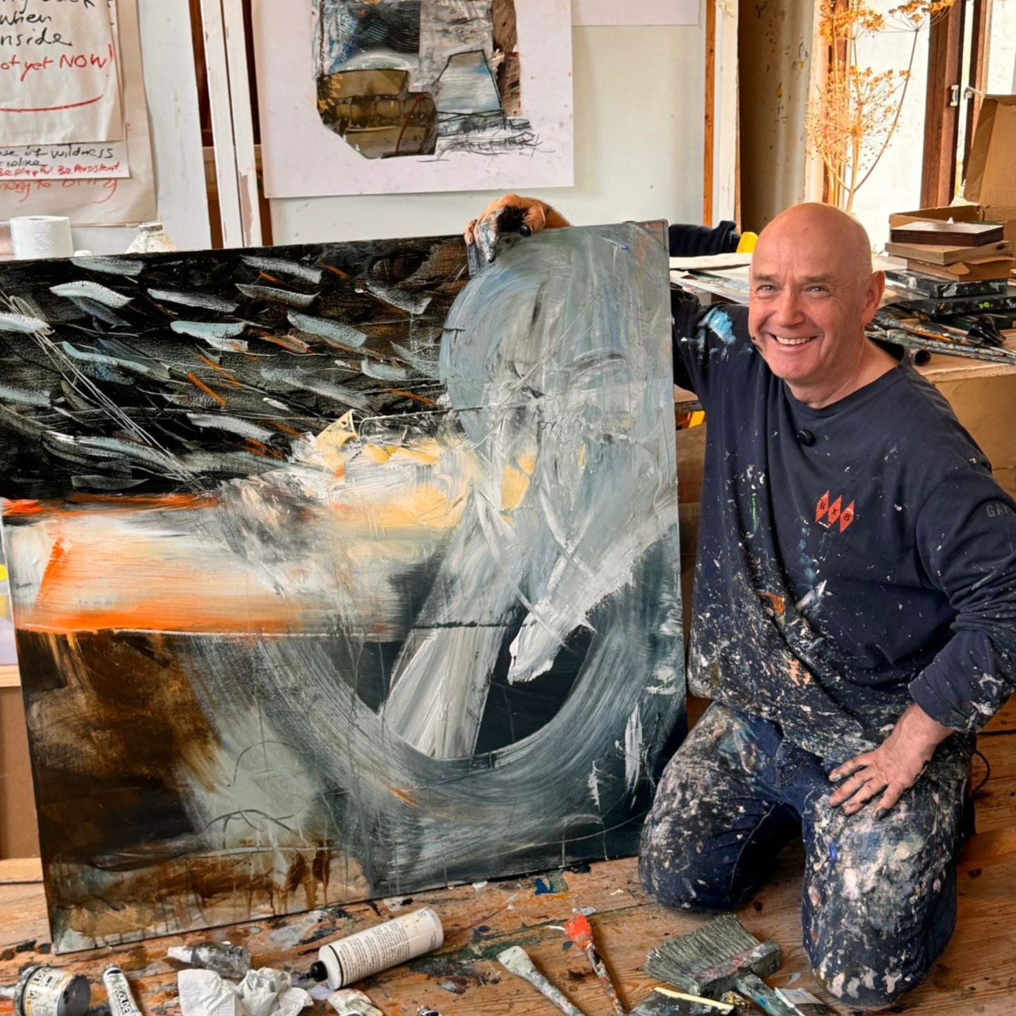 Photo of David kneeling in his studio beside a large expressive abstract painting, smiling proudly while holding the canvas, with paint tools and materials around him.