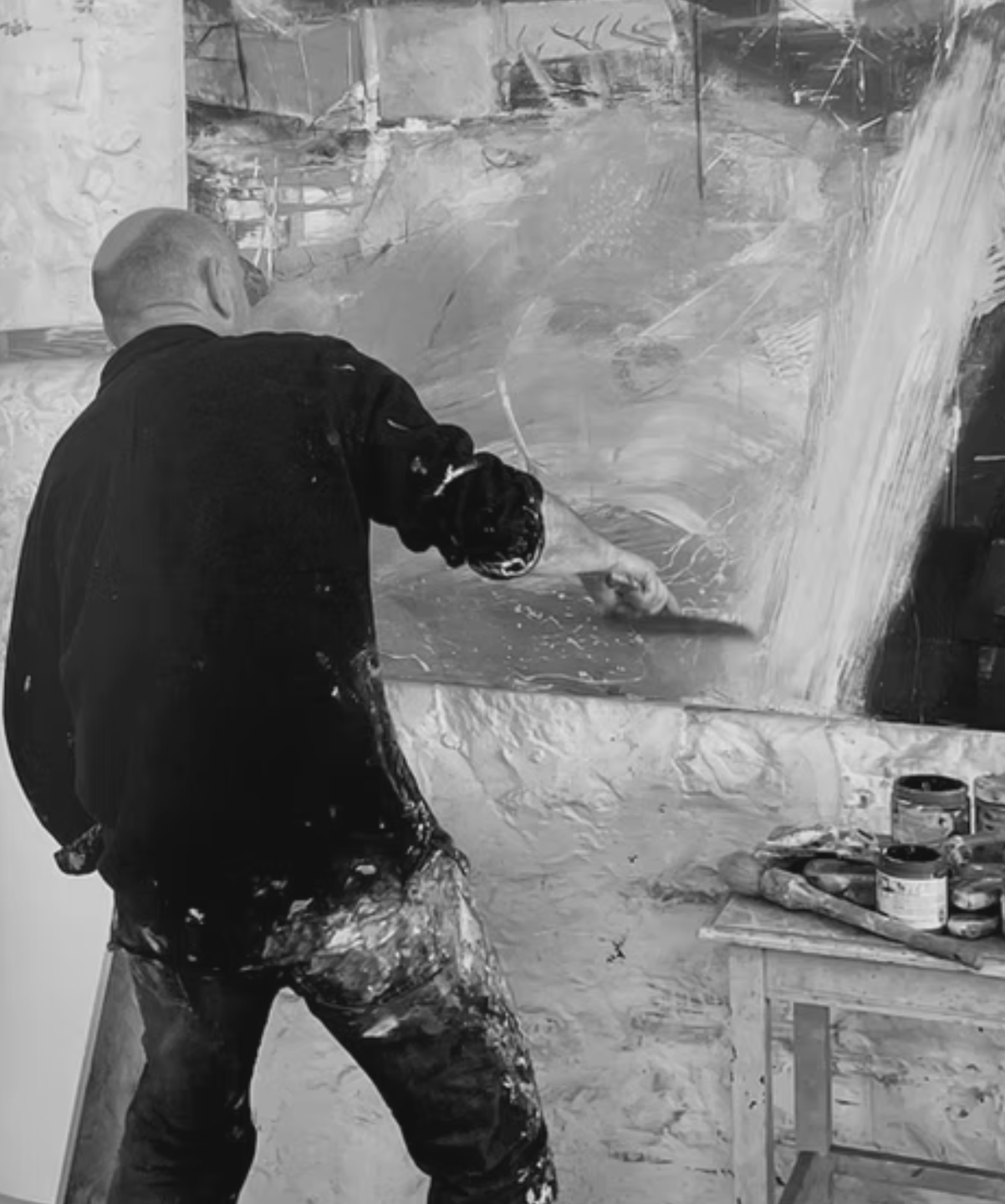 Black and white photo of David painting energetically on a large canvas in his studio, using expressive brushstrokes with paint-splattered clothing.