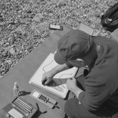 Black and white close-up of an artist’s hand sketching expressive landscape lines in a notebook, capturing the early stages of an abstract composition.