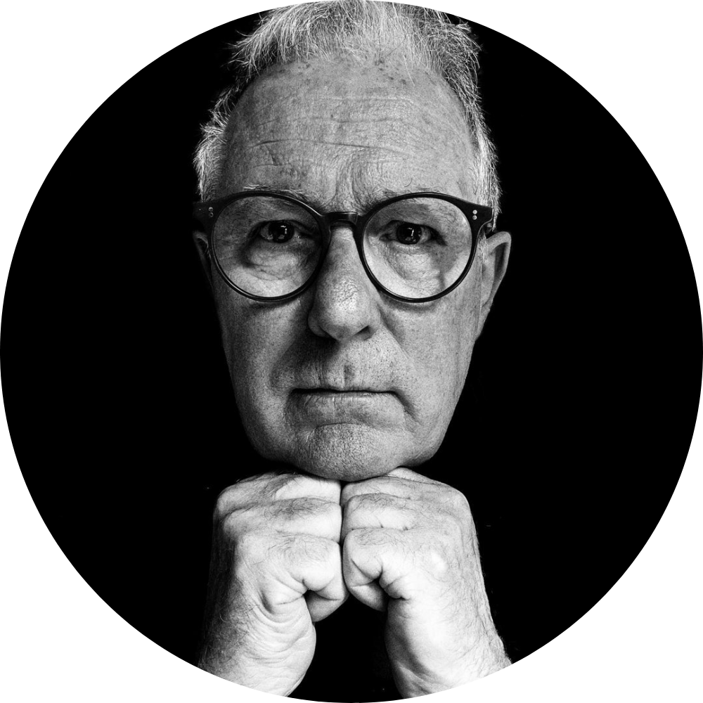 Black and white close-up portrait of a person with glasses, looking directly at the camera with their chin resting on their hands against a dark backdrop.