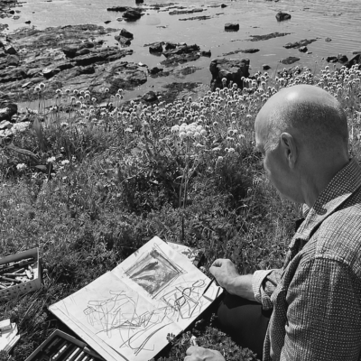 Black and white photo of David sitting on rocks outdoors, gesturing expressively as he speaks, with a sketchbook on his lap in a natural landscape setting.