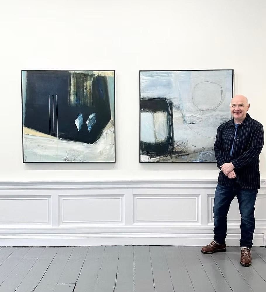 Photo of David standing in a gallery beside two large abstract paintings, smiling as he presents his work in a clean exhibition space.