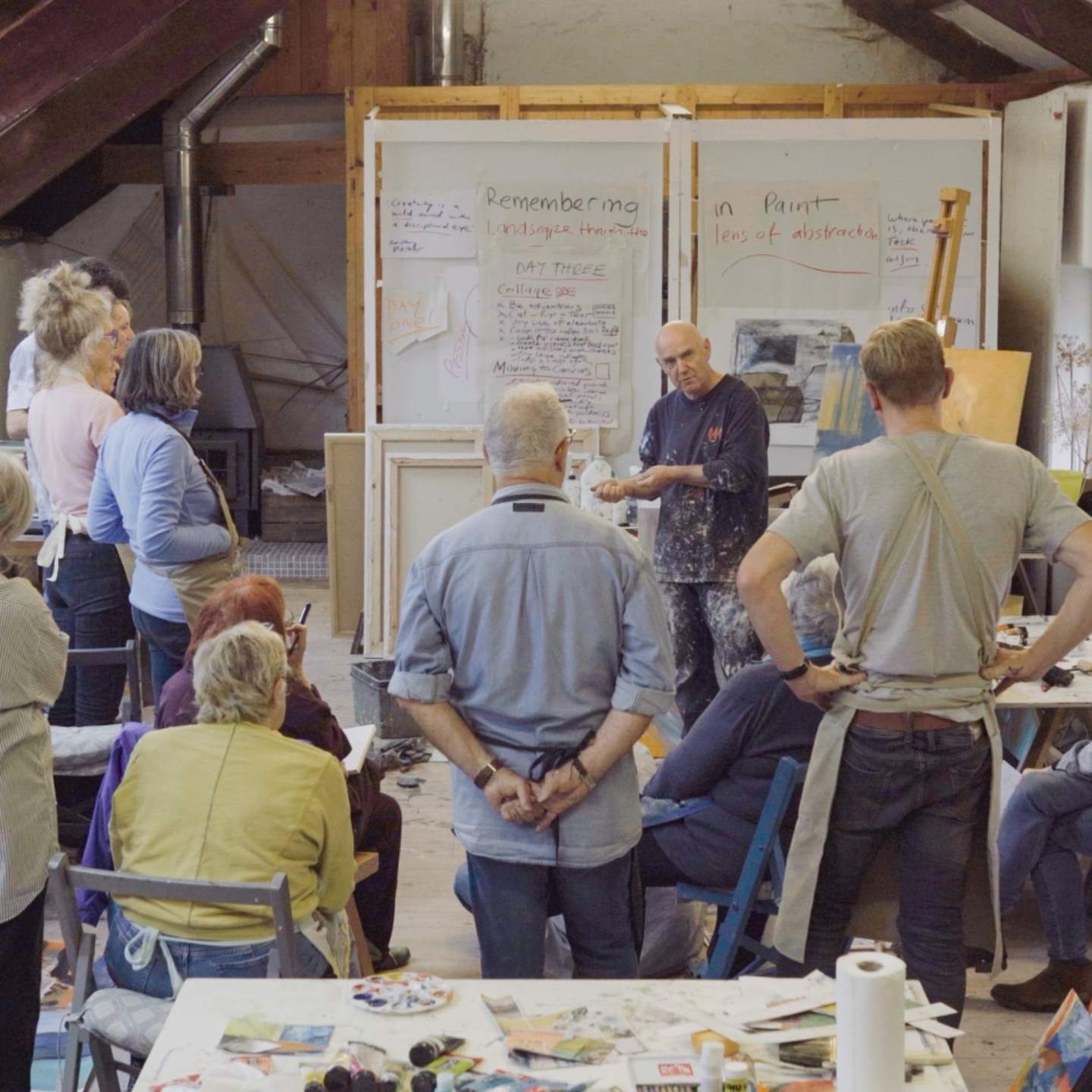 Photo of David leading a painting workshop in his studio, guiding a group of students gathered around as he explains his process in front of visual notes and artwork.