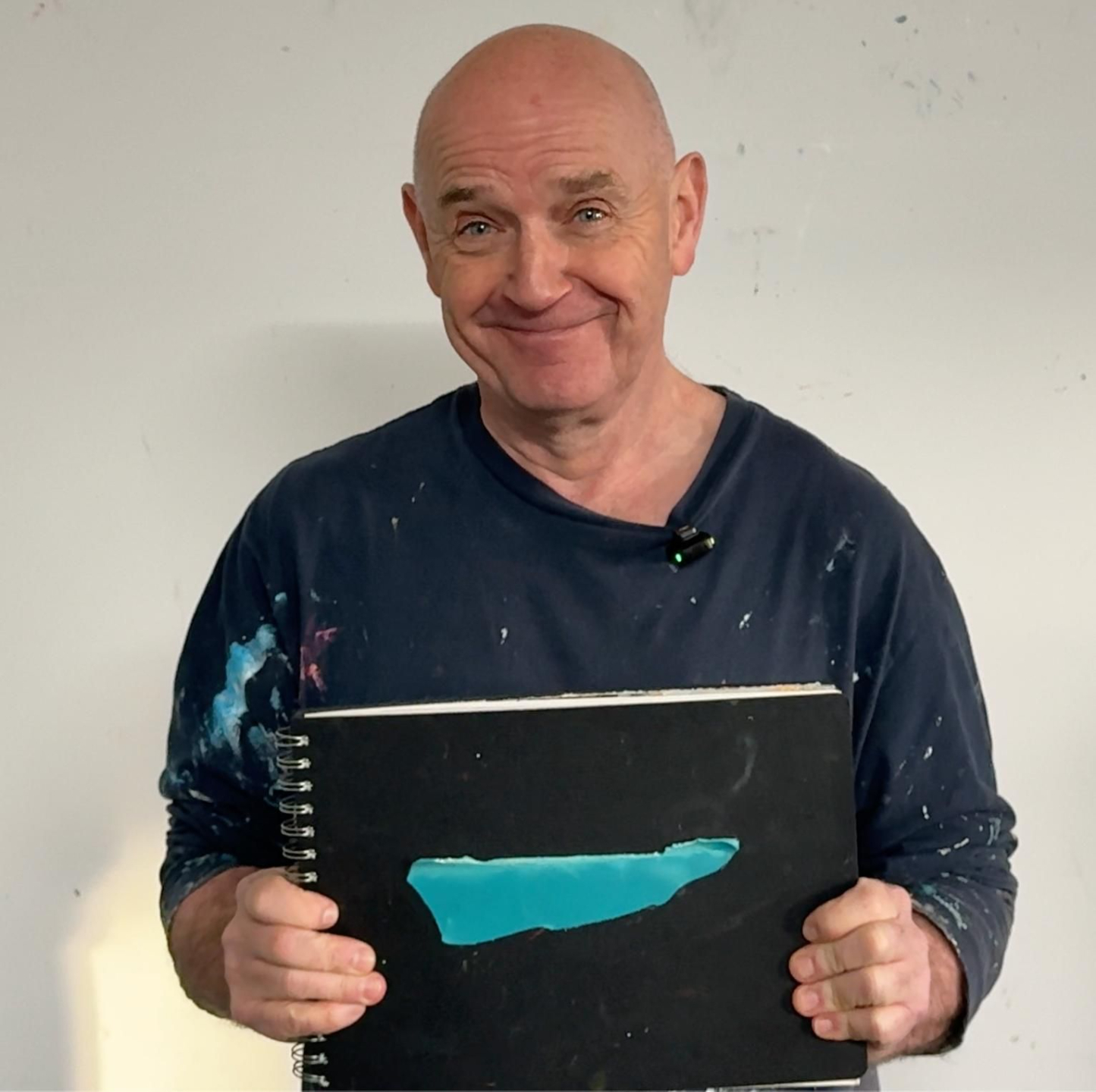 Photo of David holding a sketchbook with a bold blue painted form on the cover, smiling warmly in a simple studio setting.
