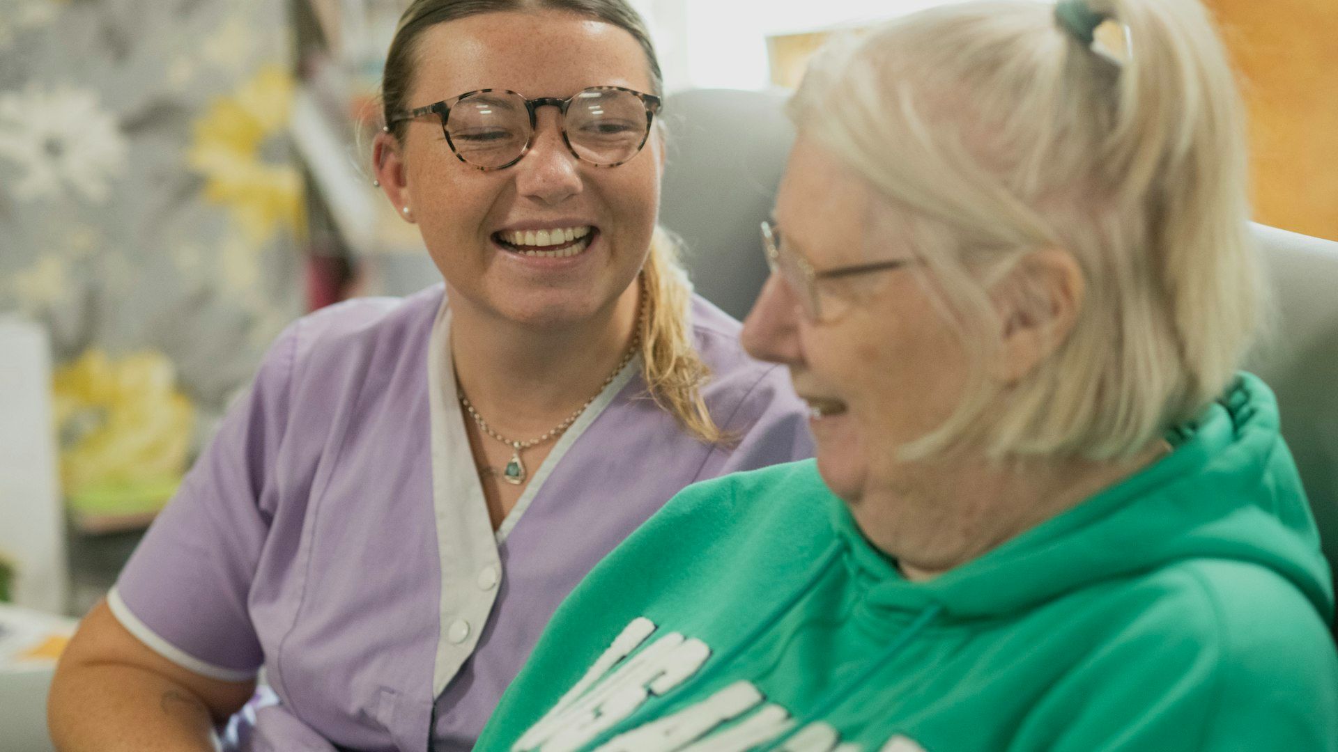 A smiling caregiver in scrubs sitting beside an elderly woman in a care home