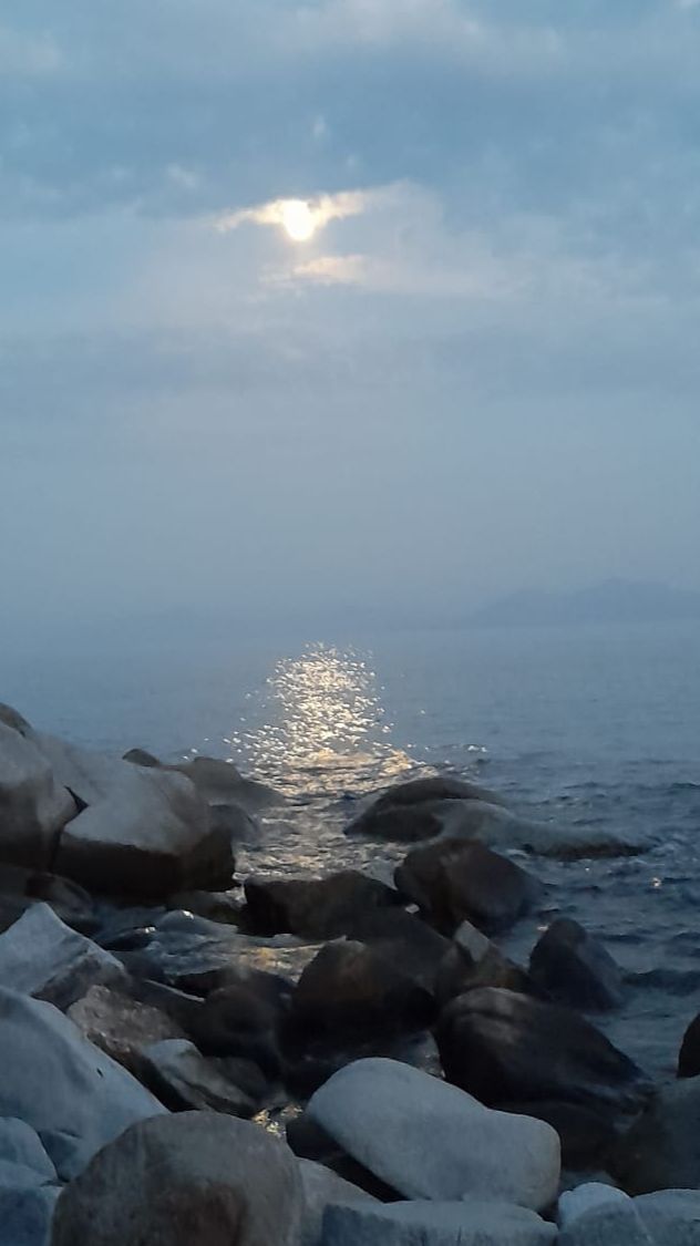 The sun on the sea through dusky clouds with dark rocks in the foreground