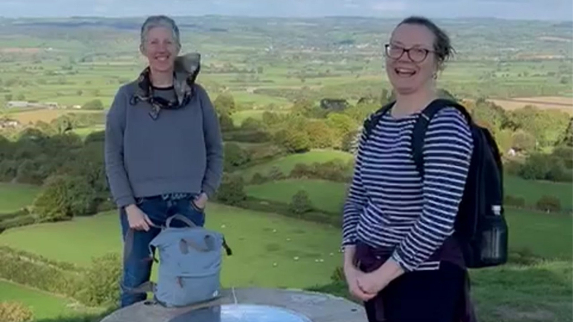2 middle-aged women smiling and laughing with a rucksack at the top of Glastonbury Tor - fields and trees behind. It's a sunny day.