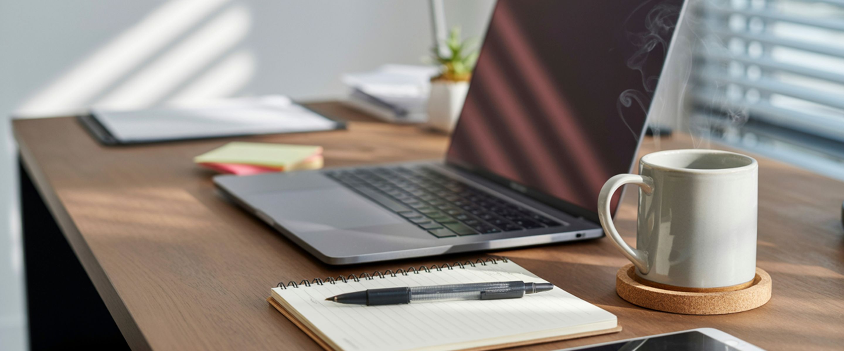 Laptop, notebook, and coffee cup sitting on a desk next to a window.