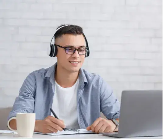 A man wearing headphones writes notes while looking at his open laptop screen.